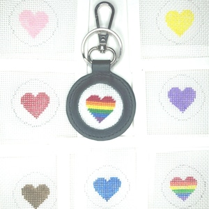 May include: A rainbow heart keychain with a black leather strap and silver key ring. The keychain is displayed with eight different colored heart designs on white canvas.
