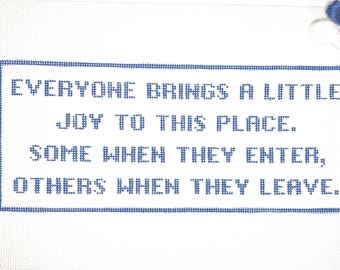 May include: A white canvas with blue cross-stitch text that reads: "EVERYONE BRINGS A LITTLE JOY TO THIS PLACE. SOME WHEN THEY ENTER, OTHERS WHEN THEY LEAVE." Blue embroidery floss is in the upper right corner.