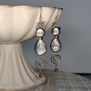 May include: A pair of dangle earrings featuring cat-shaped iridescent shell beads in gray and white tones. Each earring has a gold-toned wire spiral tail and a small dangling crystal bead at the bottom, suspended from gold-toned ear wires.