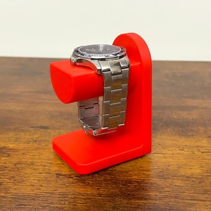 May include: A red watch stand with a silver wristwatch. The stand is a solid, angular design with a rectangular base and a curved top to hold the watch. The watch has a silver band and a dark-colored face.