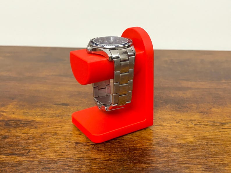 May include: A red watch stand with a silver wristwatch. The stand is a solid, angular design with a rectangular base and a curved top to hold the watch. The watch has a silver band and a dark-colored face.