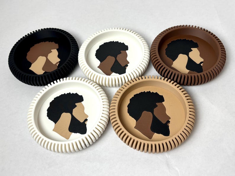May include: Five circular coasters with a ribbed outer edge, arranged in a group. Each coaster features a central graphic illustration of a person's profile with a dark beard and textured hair, rendered in varying color combinations of black, white, and brown.