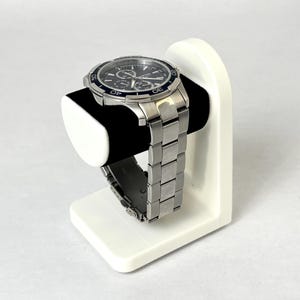May include: A white watch stand with a silver watch with a black face and blue accents. The watch is on a white stand with a black cushion.