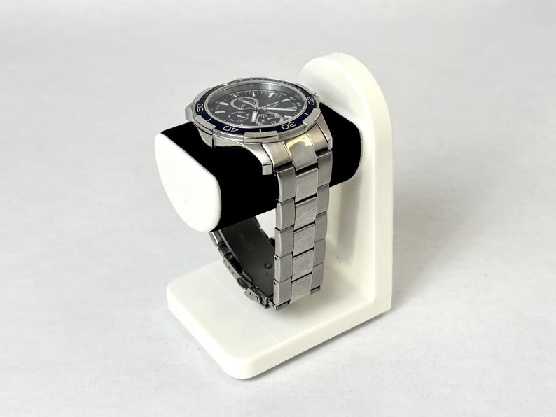 May include: A white watch stand with a silver watch with a black face and blue accents. The watch is on a white stand with a black cushion.