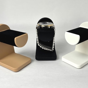 May include: Three watch display stands in beige, black and white. Each stand has a curved, padded arm to hold a watch.