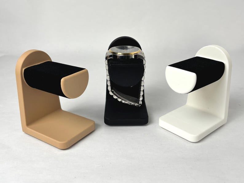 May include: Three watch display stands in beige, black and white. Each stand has a curved, padded arm to hold a watch.