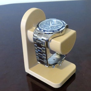 May include: A silver wristwatch with a black and blue face rests on a beige watch stand. The watch has a stainless steel band and a detailed dial. The stand is a simple, curved design, providing a display for the watch.
