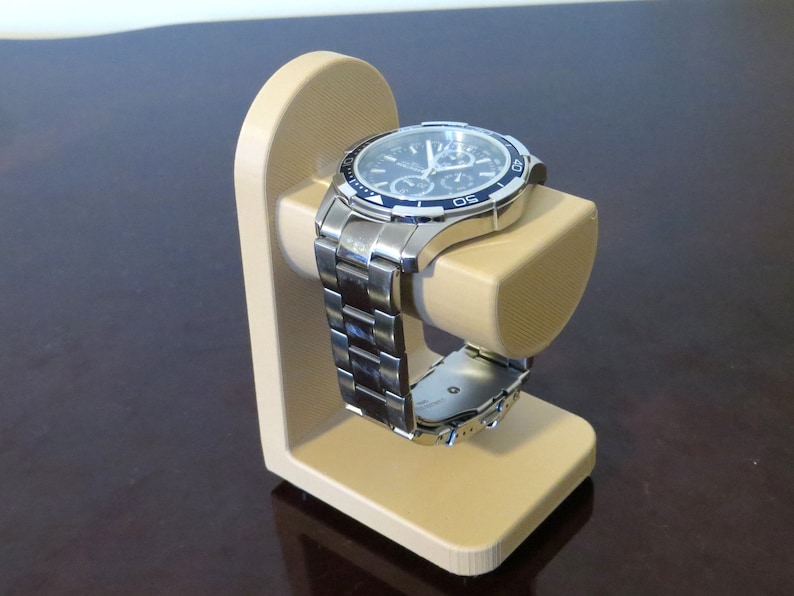 May include: A silver wristwatch with a black and blue face rests on a beige watch stand. The watch has a stainless steel band and a detailed dial. The stand is a simple, curved design, providing a display for the watch.