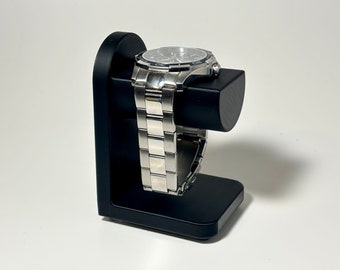 May include: A silver wristwatch with a black face is displayed on a black watch stand. The watch has a stainless steel band and is resting on a rectangular base with a curved support.