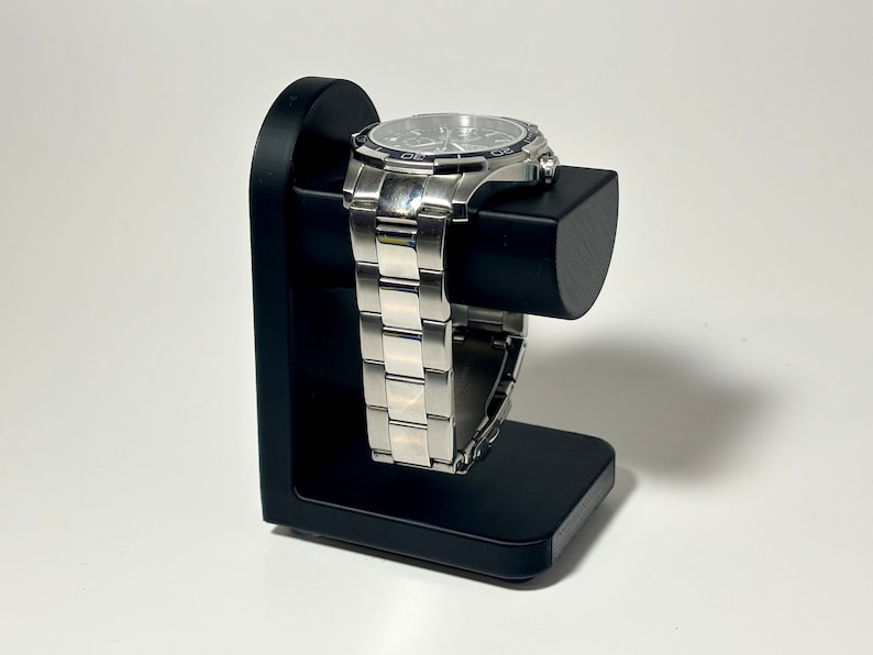 May include: A silver wristwatch with a black face is displayed on a black watch stand. The watch has a stainless steel band and is resting on a rectangular base with a curved support.