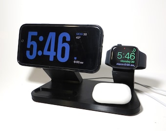 May include: A black charging station with a white wireless charging pad for AirPods. A black smartphone with a blue digital clock face displaying 5:46 and a black Apple Watch with a green digital clock face displaying 5:46 are on the charging station.