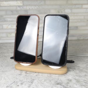 May include: Two smartphones, one with a brown case and the other with a black case, are displayed on a beige wireless charging stand. The stand is on a grey surface with a white brick background.