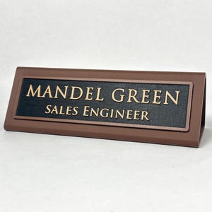 Op de afbeelding: Een bruin driehoekig naambordje voor op een bureau met een zwarte textuurinzet en gouden letters met de tekst MANDEL GREEN en SALES ENGINEER. Het naambordje heeft een stevig frame en is ontworpen om op een plat oppervlak te worden geplaatst.