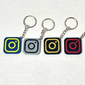 Może przedstawiać: Cztery breloczki do kluczy z logo Instagrama w różnych schematach kolorystycznych: niebiesko-żółtym, szarym, czarno-żółtym i czerwono-czarnym. Każdy breloczek ma srebrny łańcuszek i kółko.