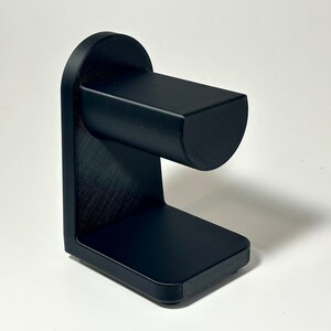 May include: A black, 3D-printed watch stand. The stand has a rectangular base, a vertical support, and a curved top section to hold a watch. The stand is made of a textured material and is designed to hold a watch.