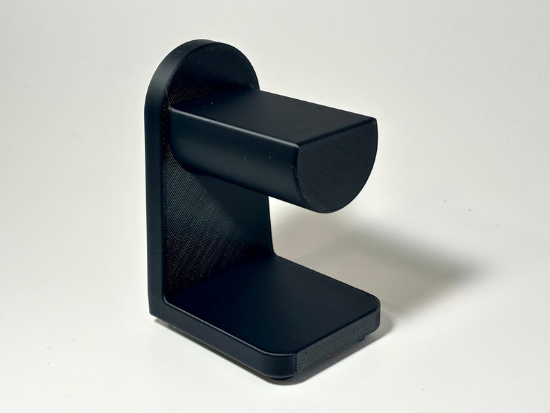 May include: A black, 3D-printed watch stand. The stand has a rectangular base, a vertical support, and a curved top section to hold a watch. The stand is made of a textured material and is designed to hold a watch.