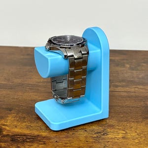 May include: A silver wristwatch with a black dial rests on a light blue watch stand. The watch has a metal band and is displayed on a stand with a curved top and a rectangular base. The stand is made of plastic.