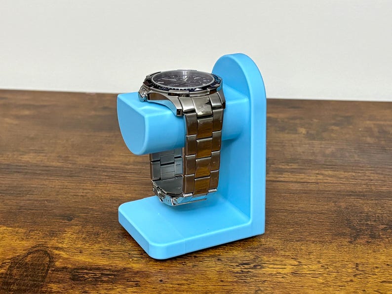May include: A silver wristwatch with a black dial rests on a light blue watch stand. The watch has a metal band and is displayed on a stand with a curved top and a rectangular base. The stand is made of plastic.