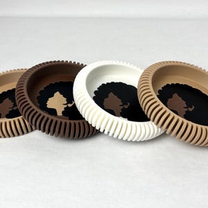 May include: Three silicone coasters with a raised rim and a textured surface. Each coaster features a silhouette of a woman with an afro hairstyle in a different skin tone: brown, white, and beige.
