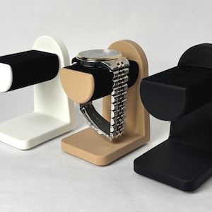 Puede incluir: Tres remontadores de relojes en blanco, beige y negro. Cada remontador tiene un cojín de terciopelo para sujetar un reloj y una base para sostener el remontador.
