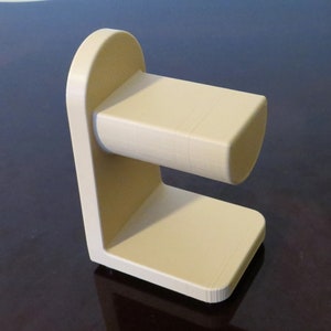 May include: A beige, 3D-printed stand with a rectangular base and a curved top. The stand features a horizontal, cylindrical component extending from the vertical support. The design is minimalist and modern, suitable for holding an item.