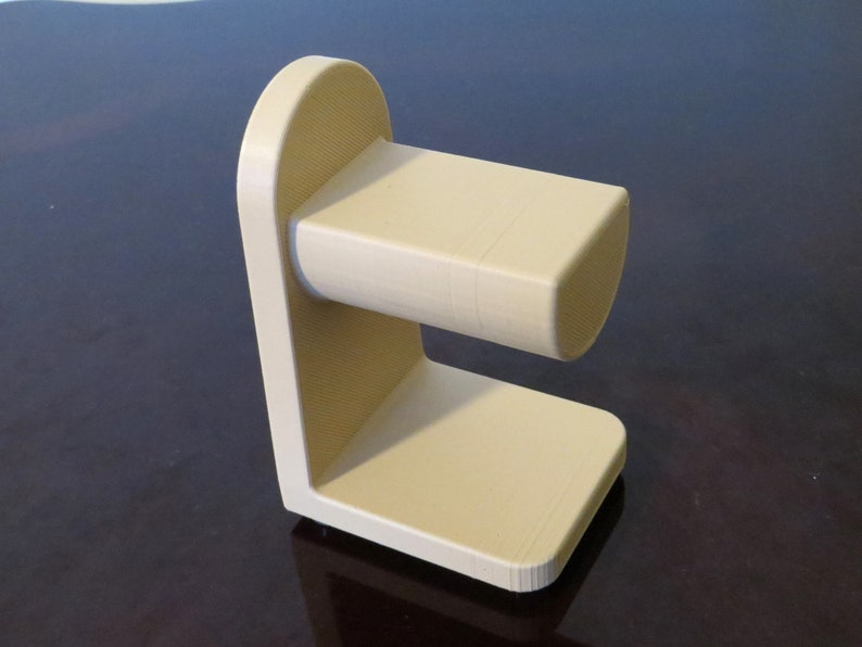 May include: A beige, 3D-printed stand with a rectangular base and a curved top. The stand features a horizontal, cylindrical component extending from the vertical support. The design is minimalist and modern, suitable for holding an item.