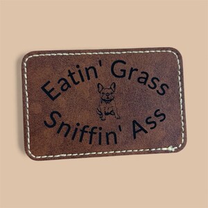 Op de afbeelding: Een rechthoekige bruine leren patch met witte stiksels. De patch heeft de tekst "Eatin' Grass, Sniffin' Ass" in zwarte letters en een kleine illustratie van een Franse Bulldog in het midden.