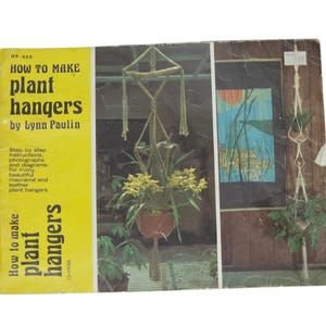 May include: A rectangular instructional book titled How to Make Plant Hangers by Lynn Paulin. The cover features a bright yellow left panel with black text and a photographic right panel displaying macrame plant hangers with green plants.