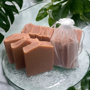 以下が含まれることがあります： 明るい茶色の手作り石鹸が透明なガラス皿に並んでいます。いくつかの石鹸はバラバラに、その他は白いシースルーの袋に入っています。緑の葉が自然な背景となり、石鹸のプレゼンテーションを際立たせています。