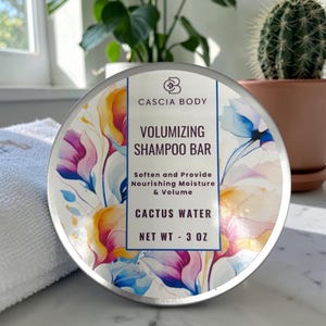 May include: A round, silver shampoo bar container with floral artwork in pink, yellow, and blue. The label reads "CASCIA BODY VOLUMIZING SHAMPOO BAR CACTUS WATER NET WT - 3 OZ". The container sits on a white surface next to a white towel.