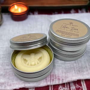 May include: Two silver tins containing a solid lotion bar. The lotion bar is white and has a bee design embossed on the top. The tin on the left is open and the lotion bar is visible. The tin on the right is closed and has a brown label that reads "Cascia Body Lotion Bar Cocoa Butter Bliss".