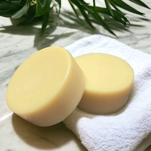 May include: Two round, light yellow soaps rest on a white, textured towel. The soaps have a smooth surface and a slightly curved shape. Green leaves are visible in the background, suggesting a natural theme.