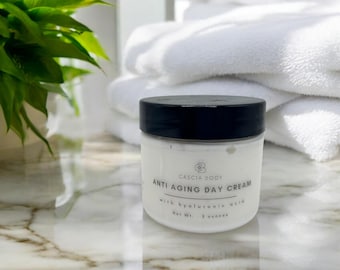 May include: A white jar with a black lid labeled "Anti Aging Day Cream" with "Cascia Body" branding. The jar contains a white cream and is set against a backdrop of white towels and a marble surface. The jar contains 2 ounces of cream.