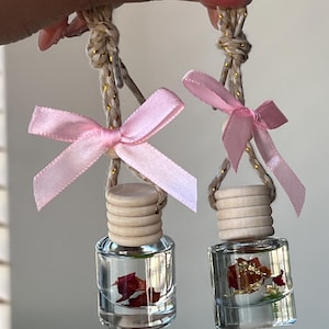 May include: Two clear glass bottles with pink bows and wooden stoppers hanging from twine. The bottles contain dried rose petals and gold flakes.