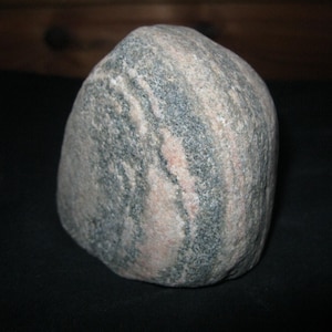 Könnte beinhalten: Ein glatter, abgerundeter Stein mit einem natürlichen Muster aus abwechselnden Bändern in Altrosa, Hellgrau und Dunkelgrau. Der Stein weist eine dichte, körnige Textur mit sichtbaren mineralischen Einschlüssen auf seiner Oberfläche auf.