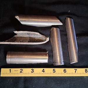 May include: Several pieces of broken metal tubing, varying in length, are scattered on a dark surface. A yellow ruler with inch markings is placed near the objects, providing scale. The metal appears to be silver in color.