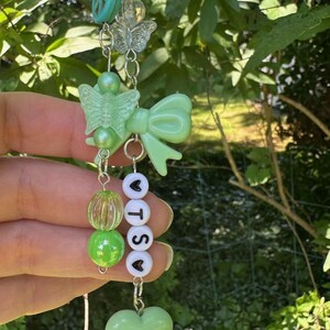 May include: A beaded charm with a silver chain, featuring a turquoise peace sign, clear butterfly, green bow, and heart-shaped beads. The charm includes beads spelling out "TSO". The charm is held up against a background of green foliage.