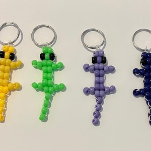 May include: Four beaded lizard keychains in yellow, green, purple, and blue. Each lizard has black bead eyes and a silver keyring. The lizards are made of small, round beads strung together to form the shape of a lizard.