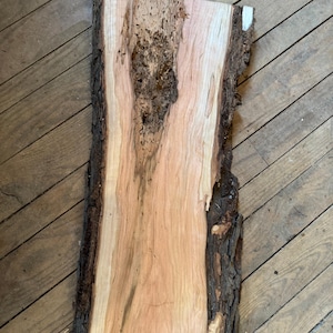 May include: A rectangular, unfinished piece of wood with a natural edge. The wood has a light brown color with darker brown and black bark along the edges. The wood grain is visible, and the surface has some natural imperfections.