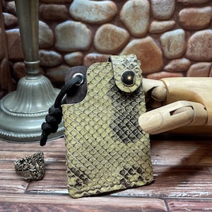 May include: A small, rectangular case with a snakeskin pattern in shades of green and brown. It has a black cord and a metal snap closure. A silver ring and a candlestick holder are in the background.