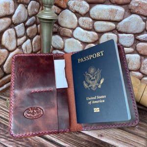 May include: A burgundy leather passport holder with a stitched border and a small pocket for cards. The holder is open to reveal a US passport inside. The holder has a logo that says "S Points Est. 2021 Leatherworks".