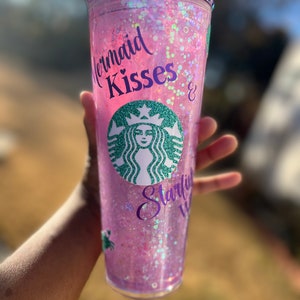 May include: A pink reusable cup with a clear lid and a glittery design. The cup has a Starbucks logo and the text "Mermaid Kisses & Starfish Wishes" in purple.