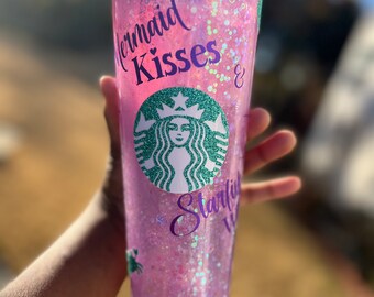 May include: A pink reusable cup with a clear lid and a glittery design. The cup has a Starbucks logo and the text "Mermaid Kisses & Starfish Wishes" in purple.