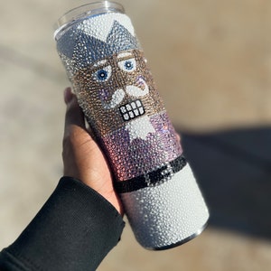 May include: A white, black, gold, and blue stainless steel tumbler with a rhinestone design of a nutcracker.