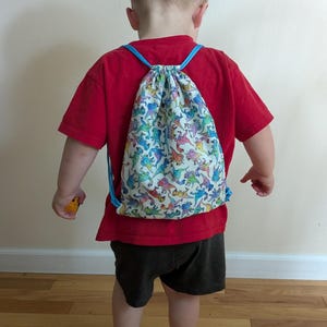 May include: A child's drawstring backpack featuring a dinosaur print. The bag has blue drawstrings and a white background with colourful dinosaur illustrations. The child is wearing a red t-shirt and black shorts. The bag is on the child's back.