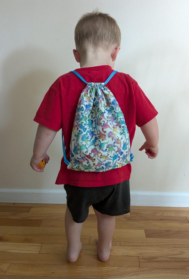 May include: A child's drawstring backpack featuring a dinosaur print. The bag has blue drawstrings and a white background with colourful dinosaur illustrations. The child is wearing a red t-shirt and black shorts. The bag is on the child's back.