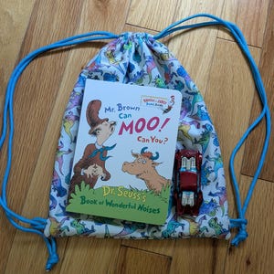May include: A drawstring bag with a dinosaur print, blue cords, and a children's book titled "Mr. Brown Can Moo! Can You?" A small red toy car is inside the bag. The book is by Dr. Seuss.