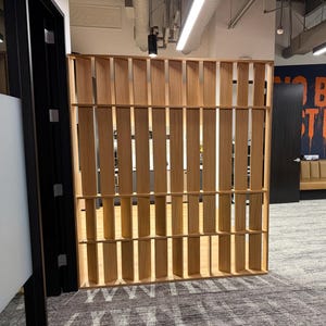 May include: A tall, rectangular room divider constructed from light-coloured wood, featuring a series of vertical slats arranged in a grid pattern. The structure is freestanding and provides a semi-transparent partition within the office space.