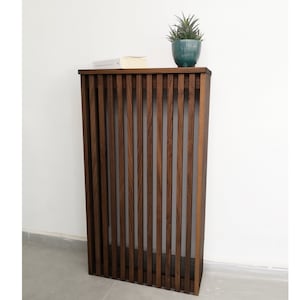 Op de afbeelding: Een donkerbruine houten consoletafel met een lattenontwerp. De tafel heeft een klein plankje en een bovenblad. Een kleine plant in een pot staat op het bovenblad.