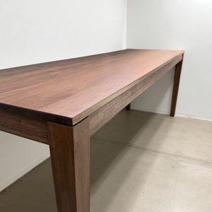 May include: A rectangular wooden table with a dark brown finish. The table has a simple design with four square legs and a smooth, flat surface. The wood grain is visible on the tabletop.